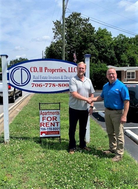 Two men shaking hands beside a real estate sign and a “For Rent” sign outdoors.