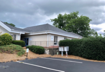 Single-story building with a ramp, hedges, and a parking lot under cloudy skies.