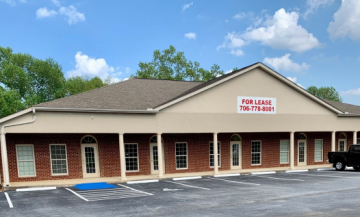 Single-story brick storefront with a “For Lease” sign and empty parking lot under a partly cloudy sky