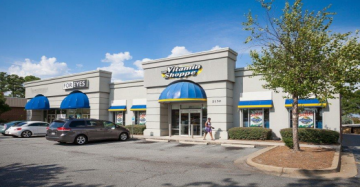 Storefront of a shopping center with blue awnings, parked cars, and a tree-lined parking lot under a sunny sky