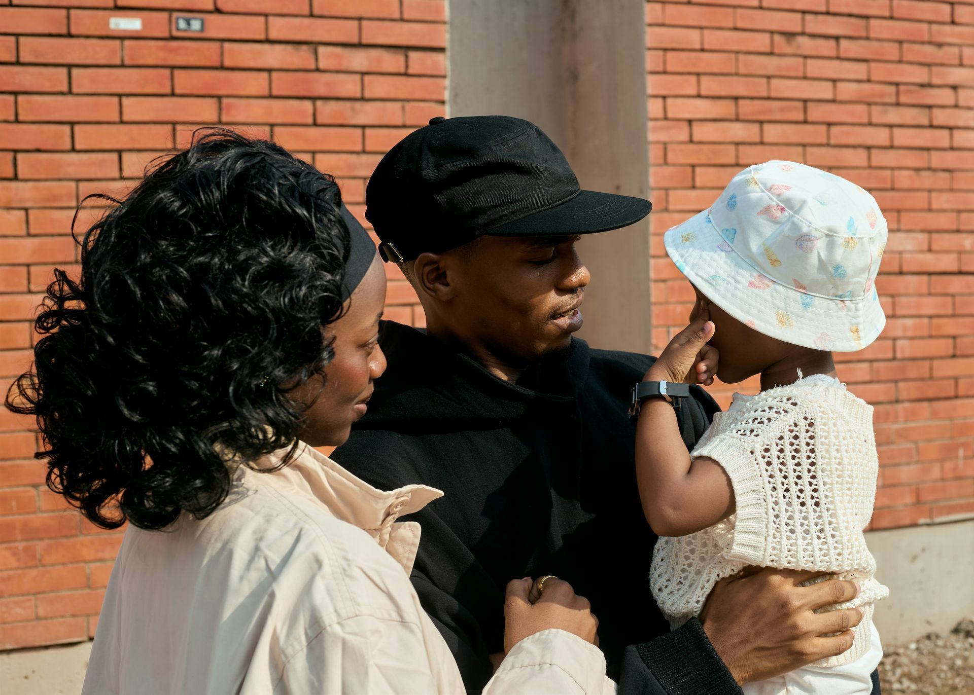 Two adults holding a child in a white hat and crochet top against a brick wall.