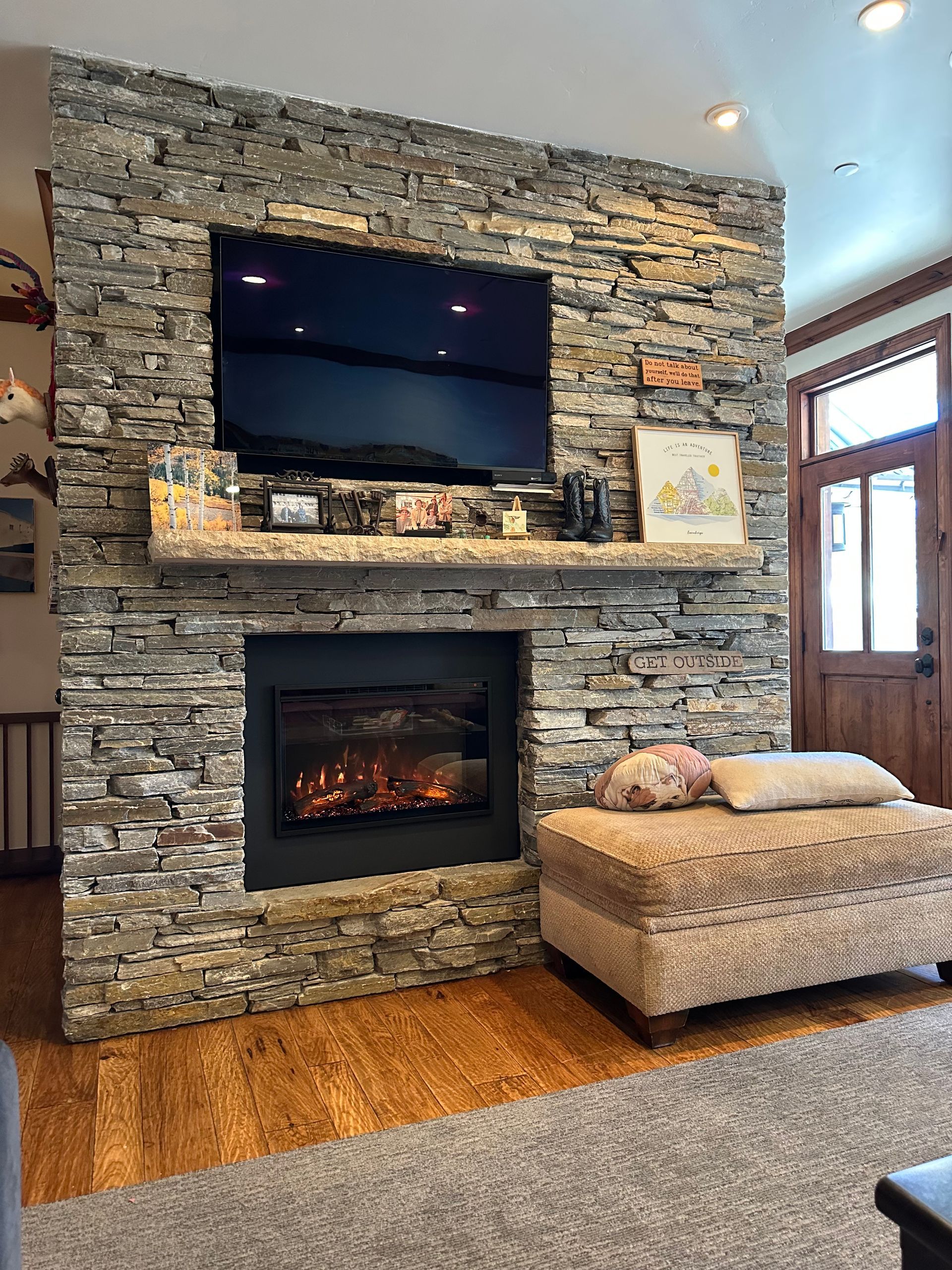 Living room fireplace with stone surround, mounted TV, woven ottoman, and wood floor