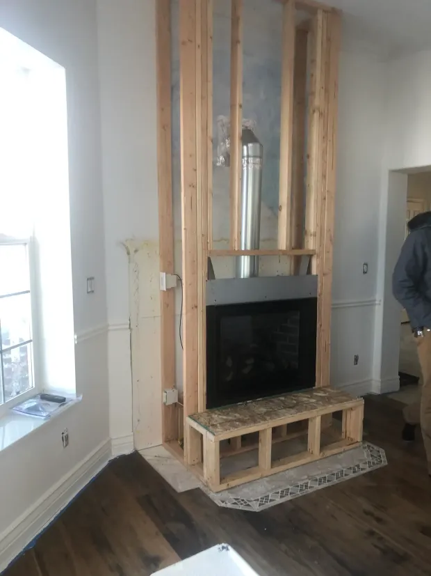 Fireplace under construction, wood frame, black insert, chimney. Man standing in room with white walls, wood floors.
