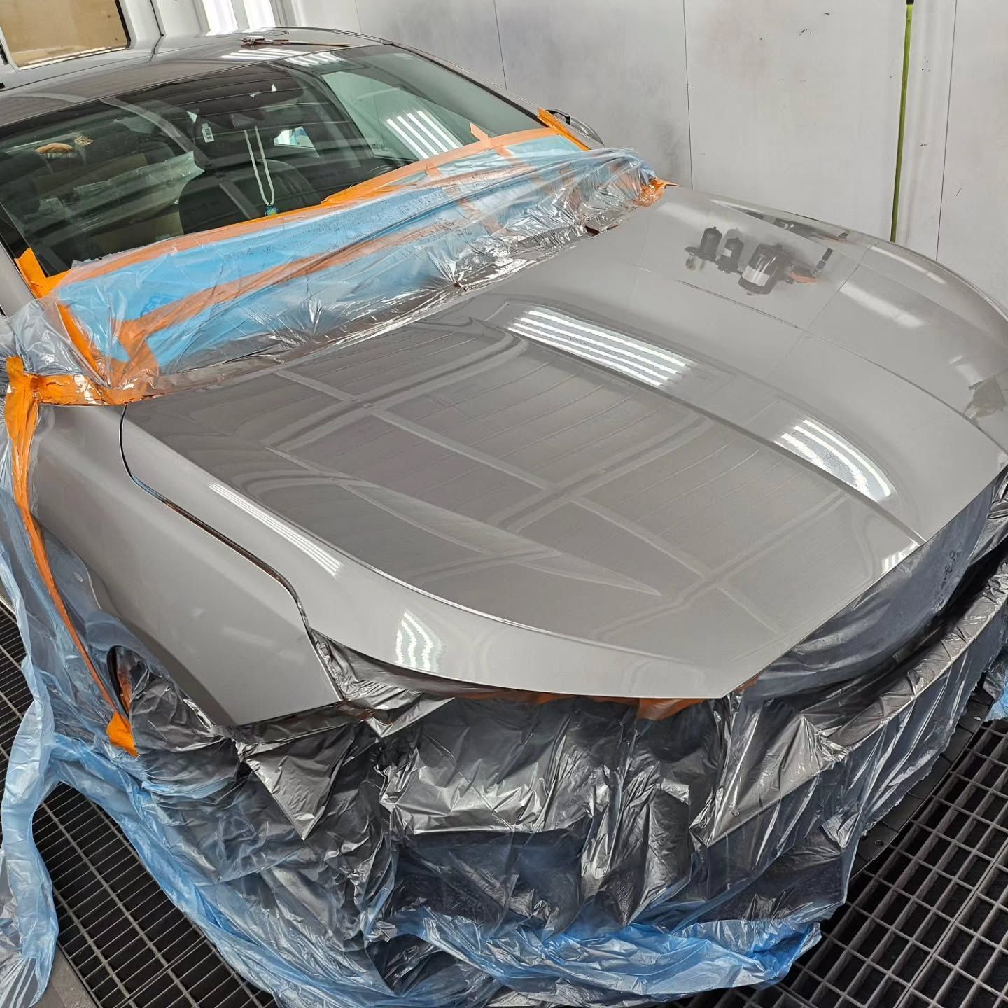 Gray car hood and windshield masked with plastic and tape in an auto body shop during painting.