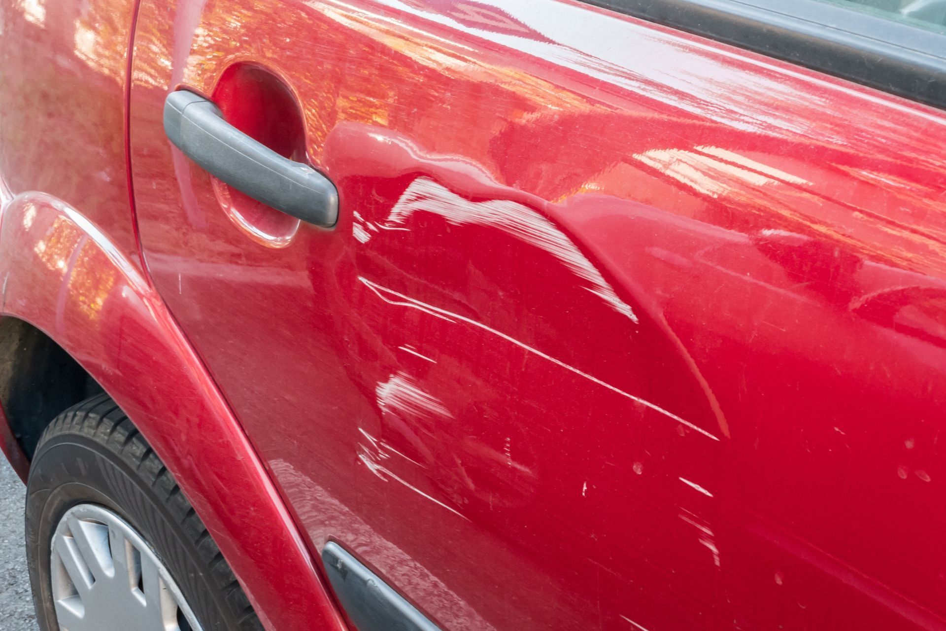 Close-up of a red car door with a noticeable dent and scratch near the handle