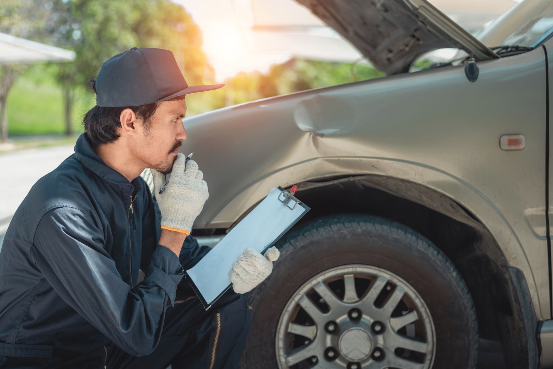 Mechanic inspecting a car’s front wheel with the hood open and a clipboard in hand
