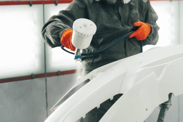Person spray-painting a white car bumper in a workshop, wearing orange gloves and a respirator.