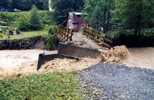 Flooded bridge, brown rushing water, red building in background.