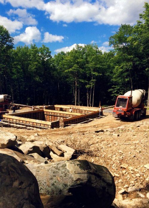 Construction site: Foundation forms ready, concrete truck. Blue sky, trees in background.