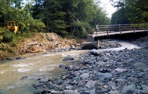 Muddy river flowing under a wooden bridge, rocky banks, trees in the background, yellow construction vehicle on the left.