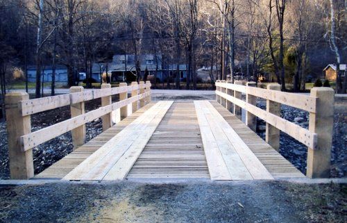 Wooden bridge with railing, spanning over a gravel area, surrounded by trees.