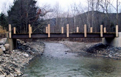 A bridge over a stream. Steel beam supported by concrete with wooden posts along the top.