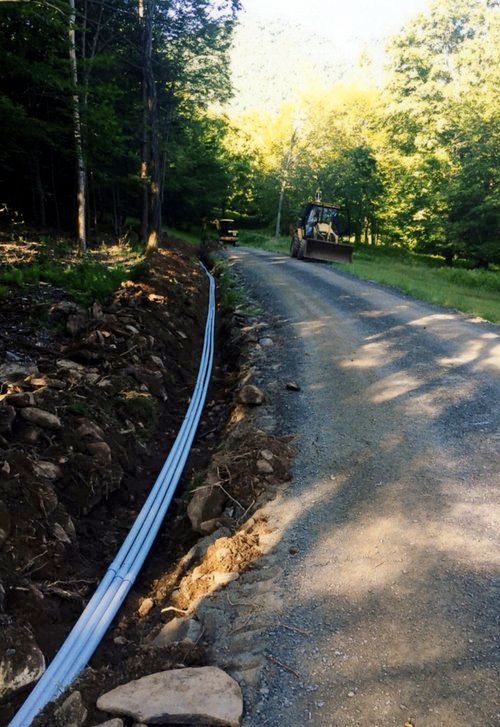 Trench with blue pipes next to a gravel road. A small excavator is visible in the distance.