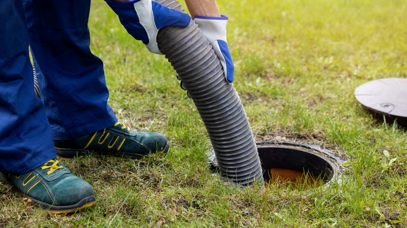 Person in blue coveralls and gloves pumping a septic tank in grassy yard.
