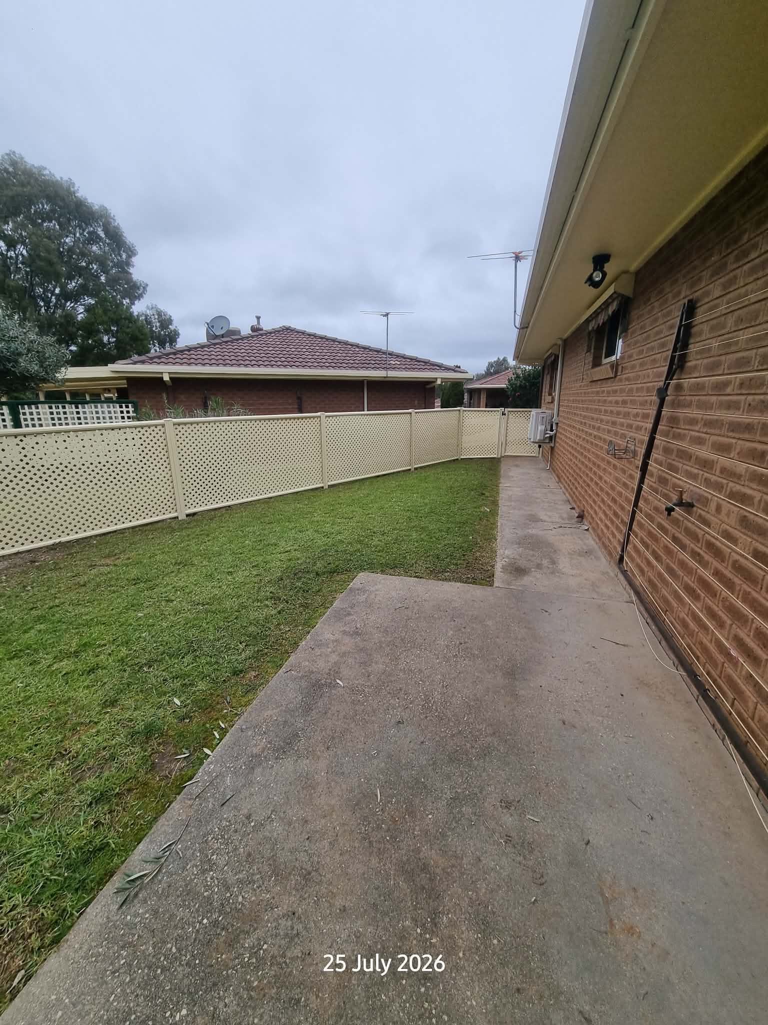 Side Yard With Brick House Wall, Concrete Walkway, Grassy Lawn, and a White Fence Under Cloudy Skies — A Plus Project Maintenance in Wodonga, VIC