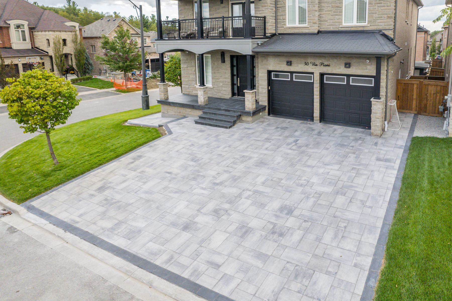 Suburban house with gray brickwork driveway, grass, and two-car garage. Black garage doors and stone facade.
