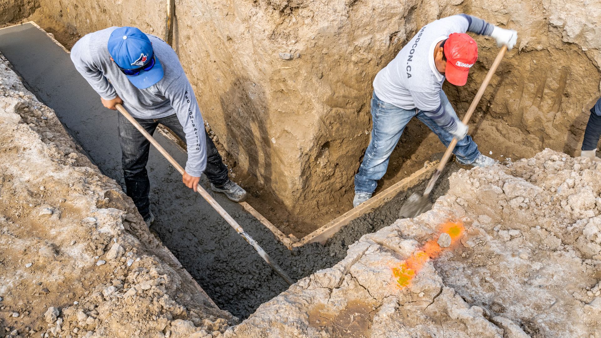 Two workers in hard hats digging a trench with shovels and laying black pipe at a construction site