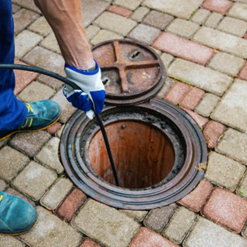 Gloved worker lowers a tool into an open manhole on a brick-paved sidewalk