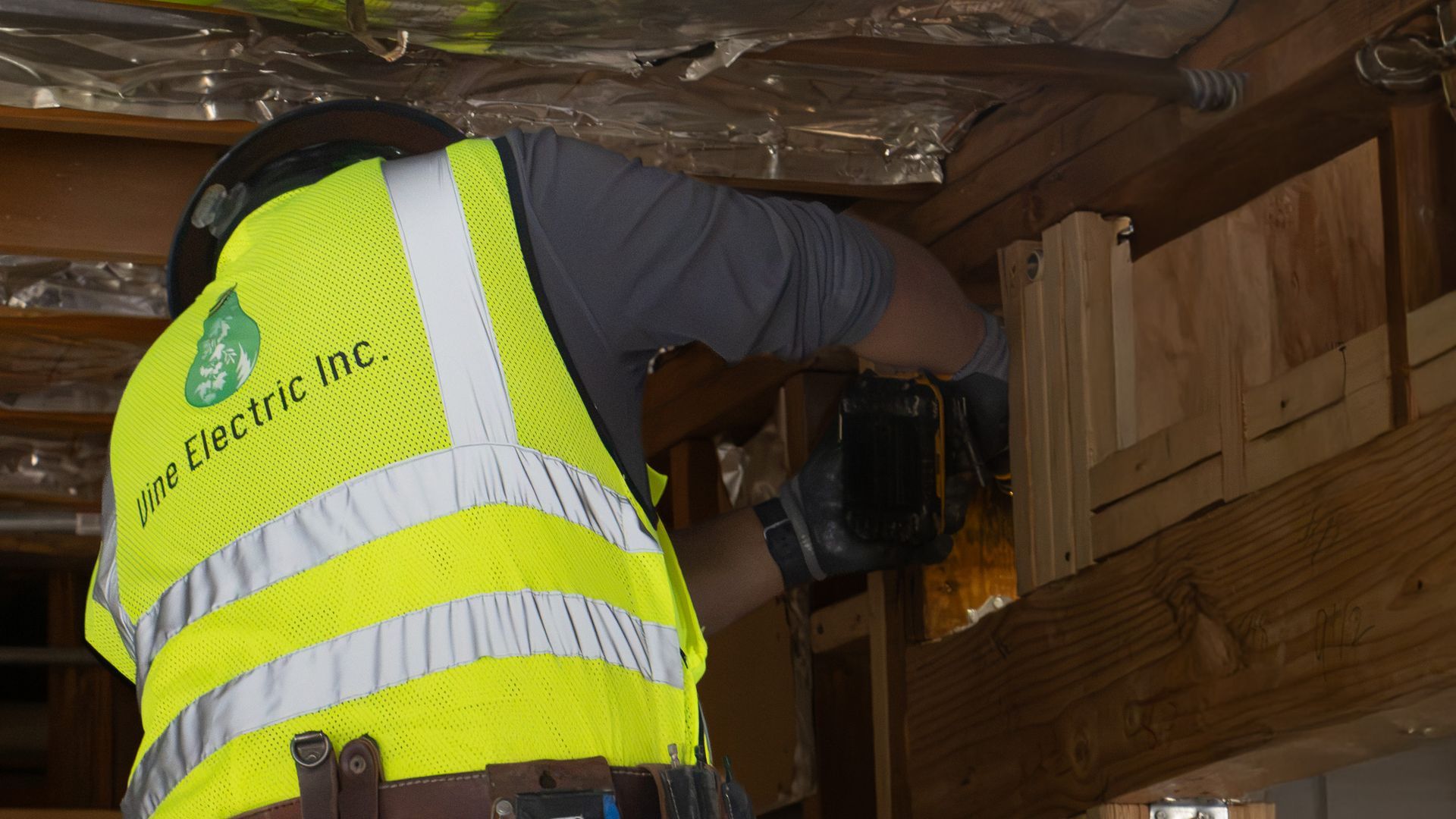 Worker in a neon yellow safety vest repairing wiring inside a wooden attic space