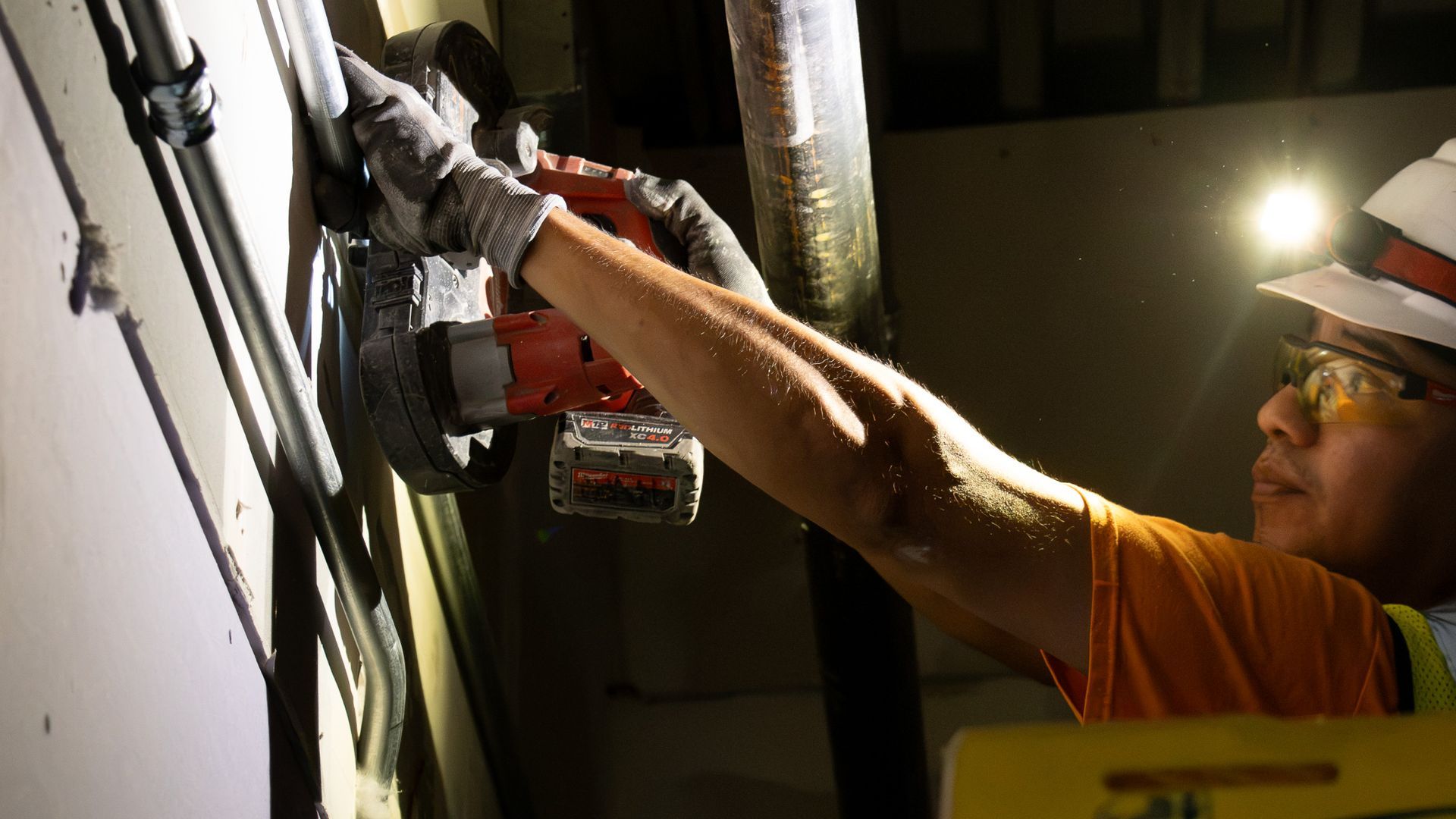 Worker in safety gear using a power tool to drill or install equipment on a wall inside a dim industrial setting