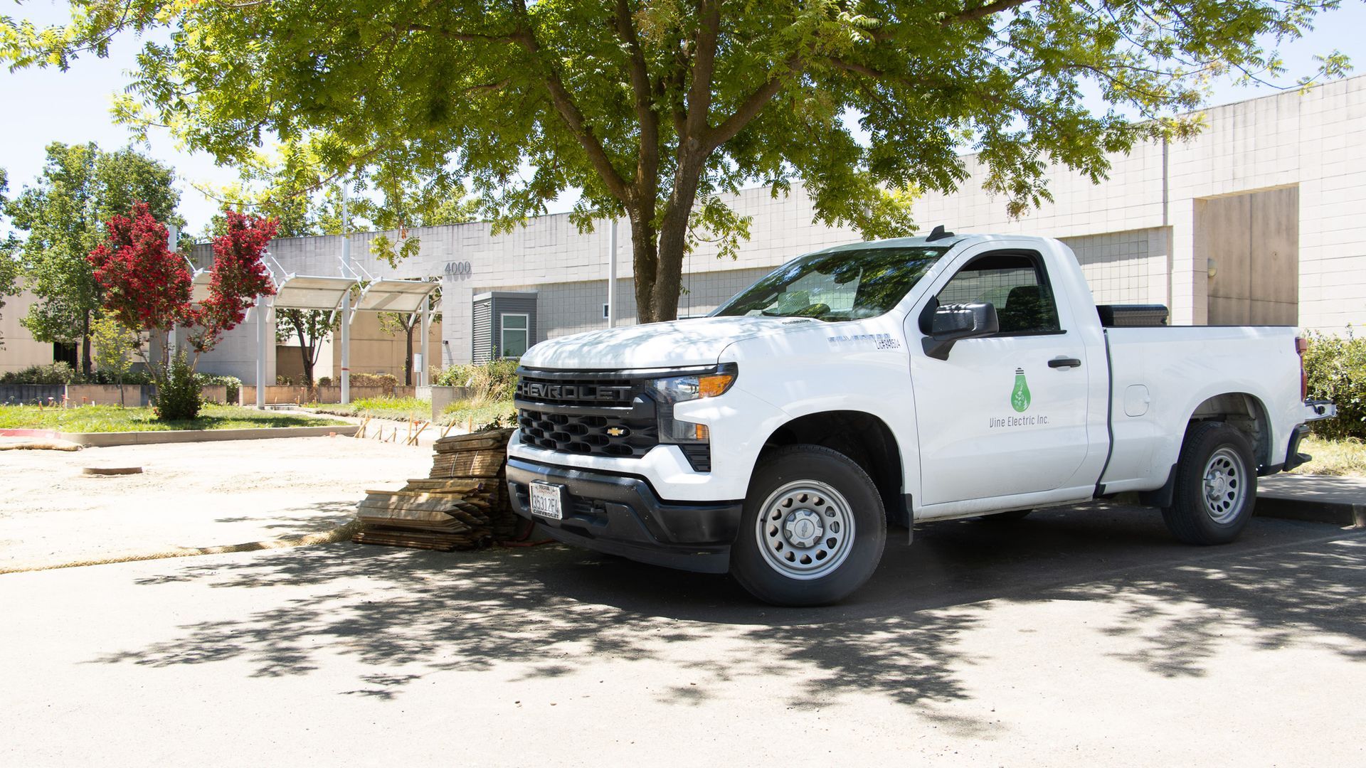White pickup truck parked in front of a building under leafy trees on a sunny day