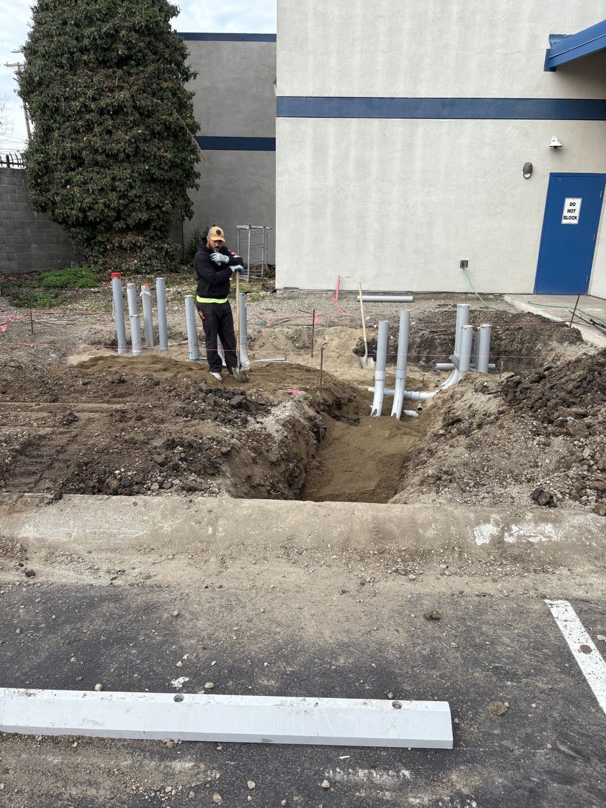 Construction site with dirt trench, metal pipes, and a person standing near a building entrance