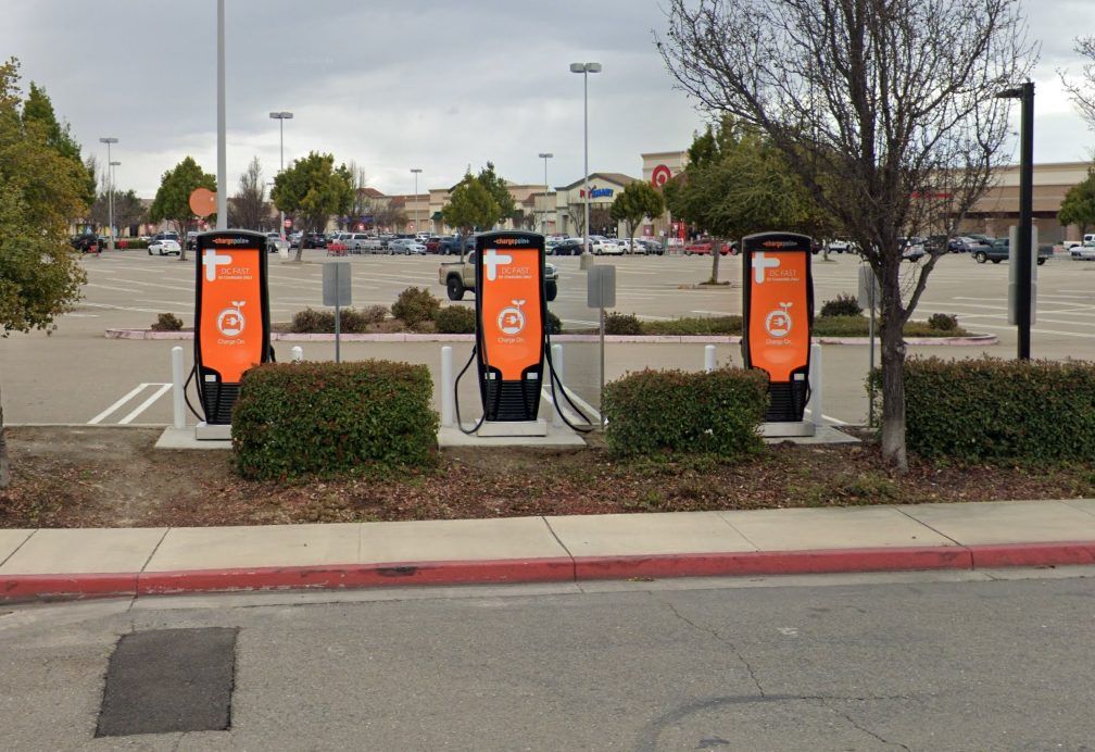 Three orange EV charging stations in a parking lot beside a curb and shrubs