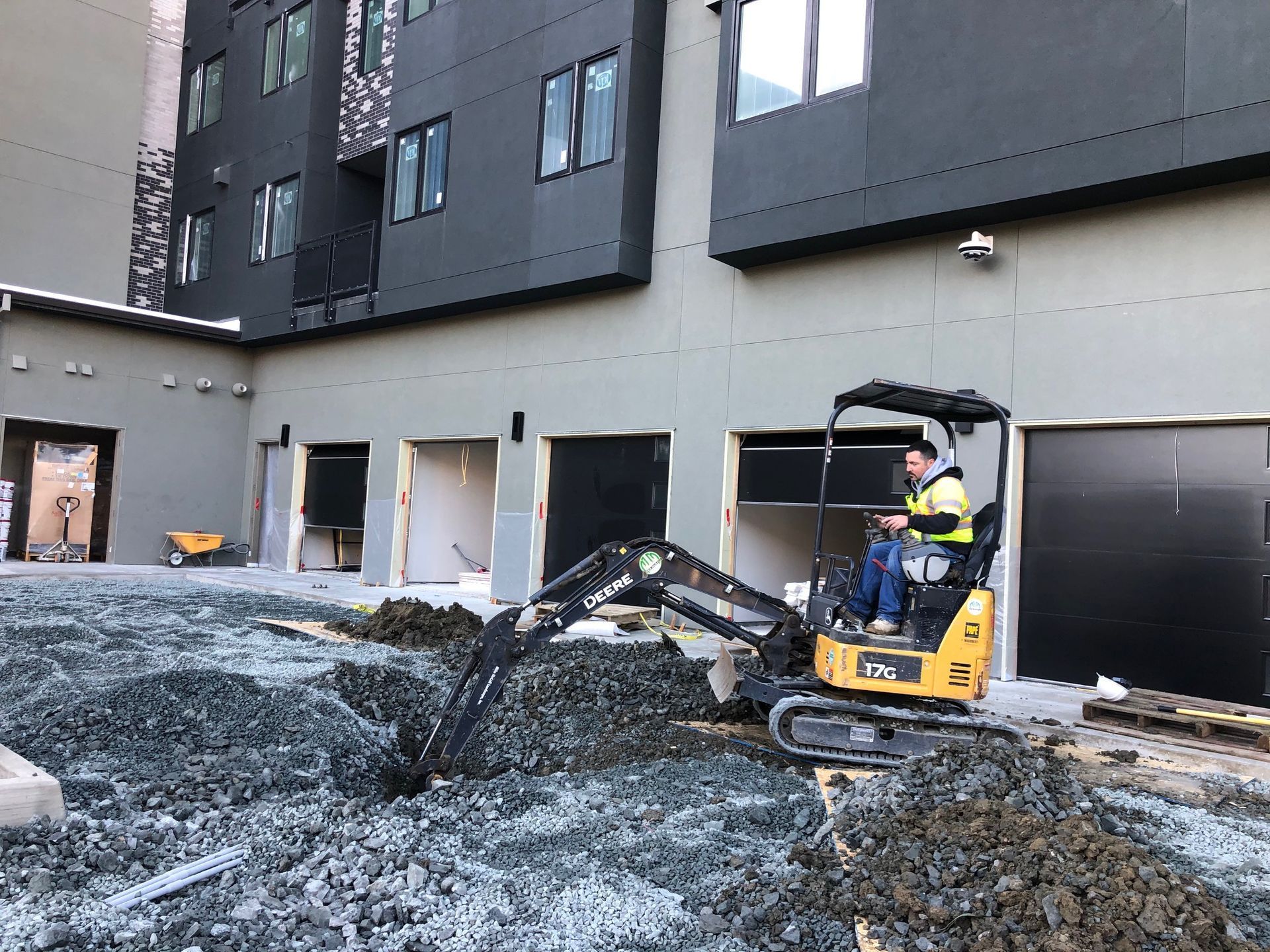 Construction worker on a yellow excavator clearing gravel beside a modern building under construction.
