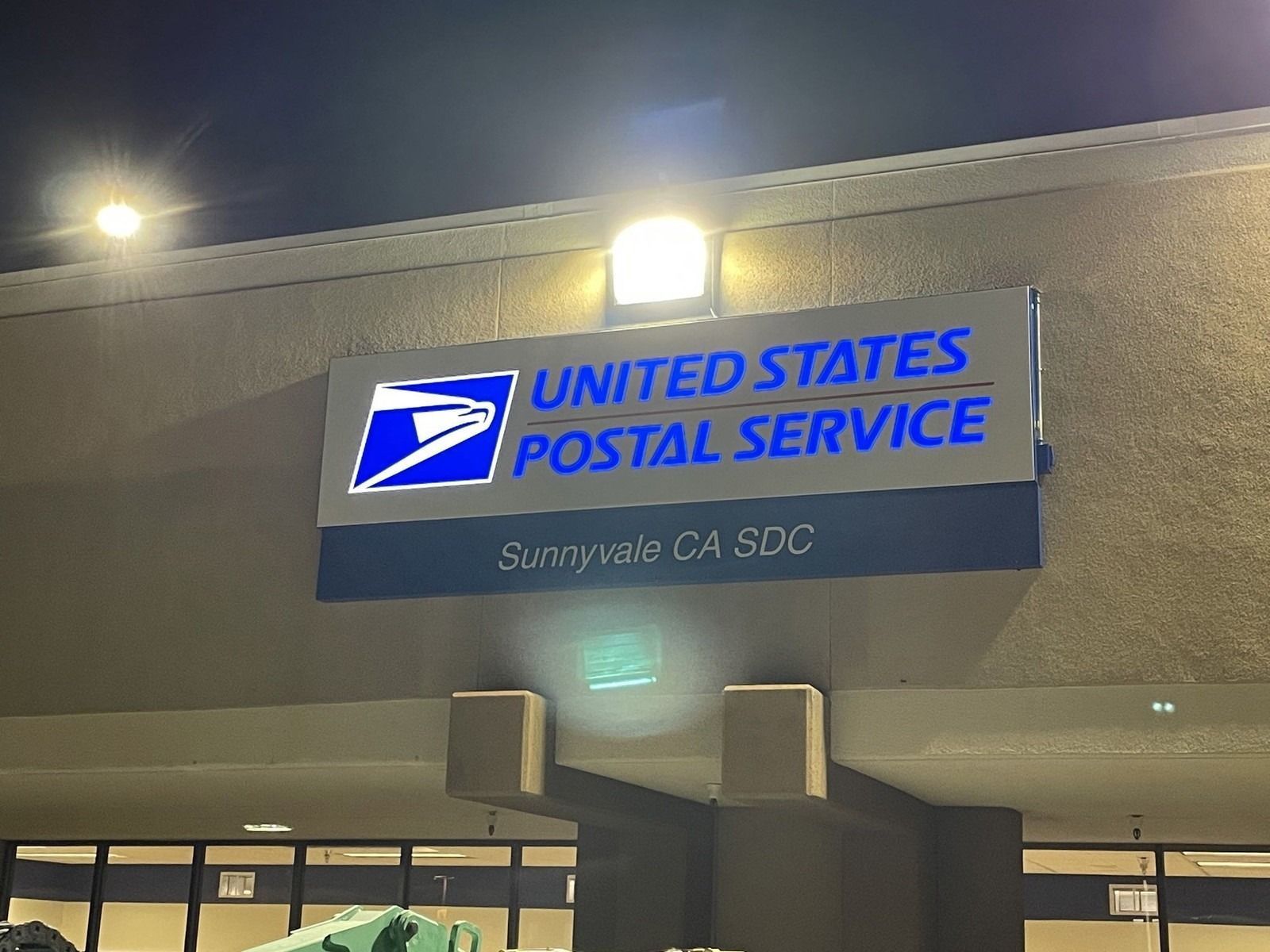 United States Postal Service sign on a building at night, lit in blue and white