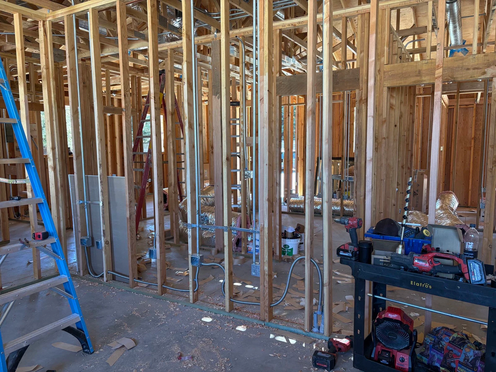 Interior of a building under construction, wooden frames with electrical wiring visible, tools on a black cart, blue ladder.