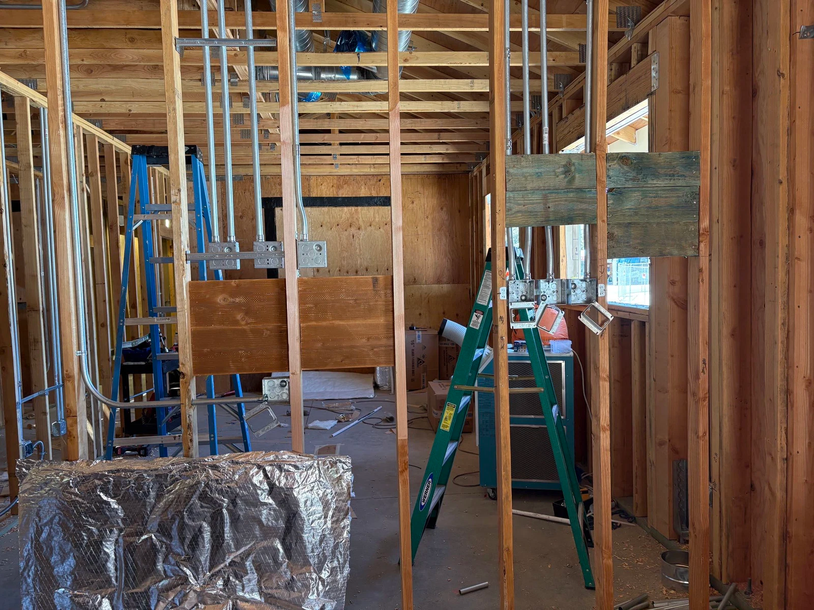 Interior of a building under construction. Studs and wiring are visible. A ladder and other tools are present.