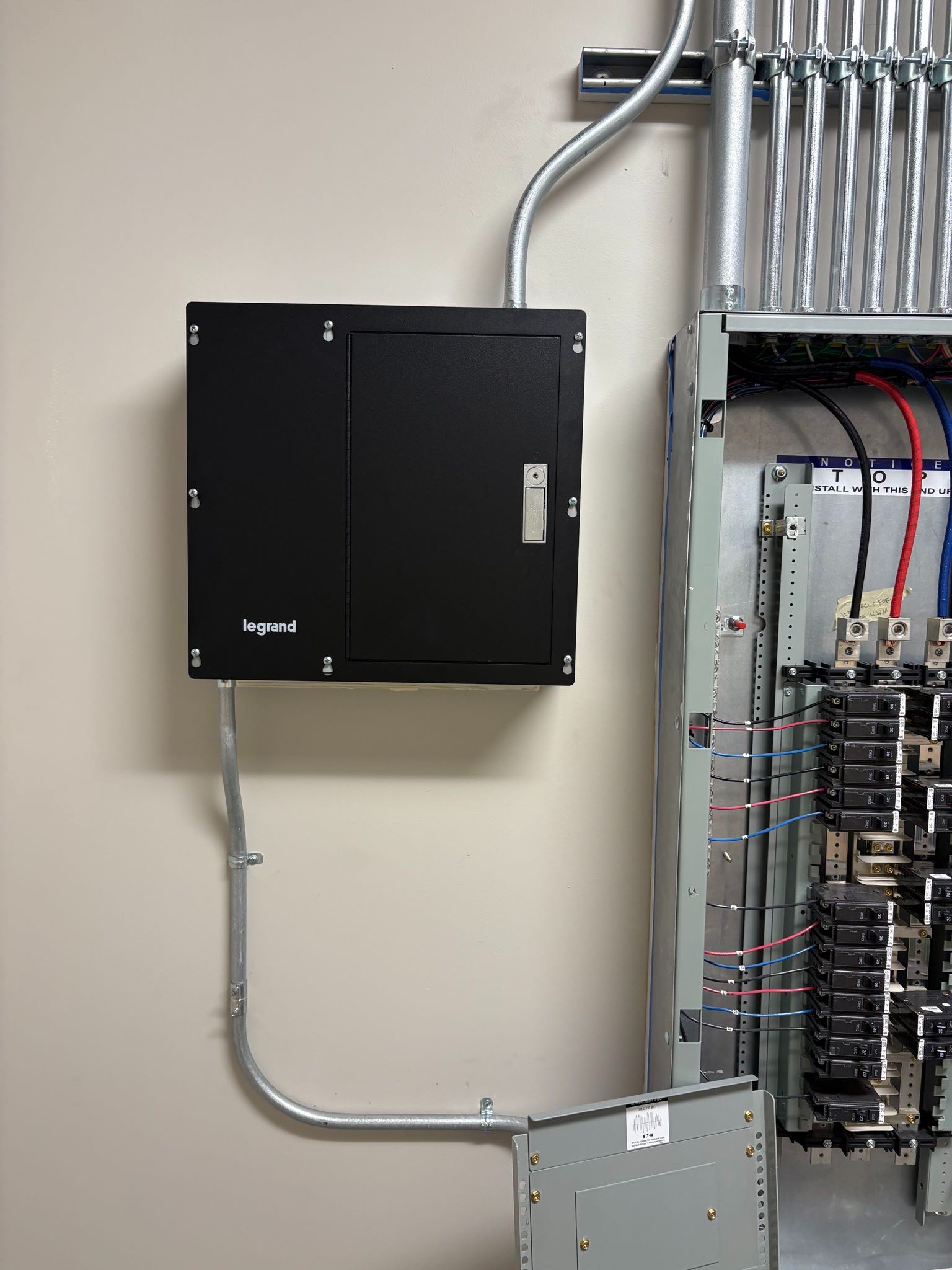 Black electrical box on a wall with conduit and a metal panel with wiring.