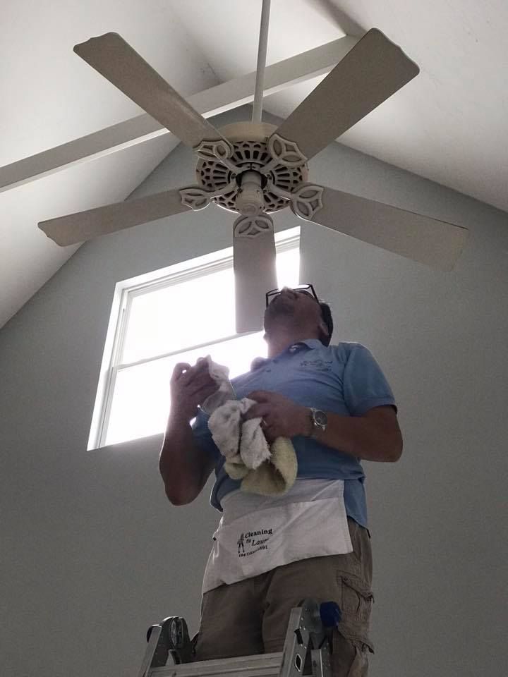 A man is standing on a ladder cleaning a ceiling fan.