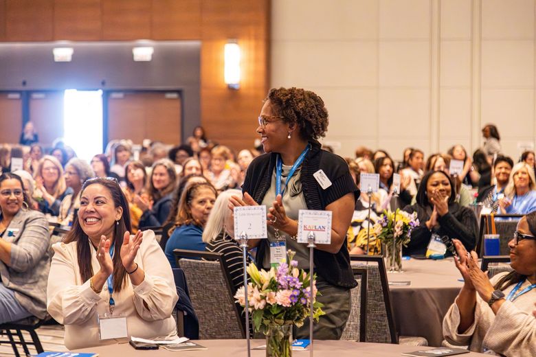 A crowd of people seated at round tables in a conference hall are clapping, with some individuals standing to cheer. A crowd of people seated at round tables in a conference hall are clapping, with some individuals standing to cheer.