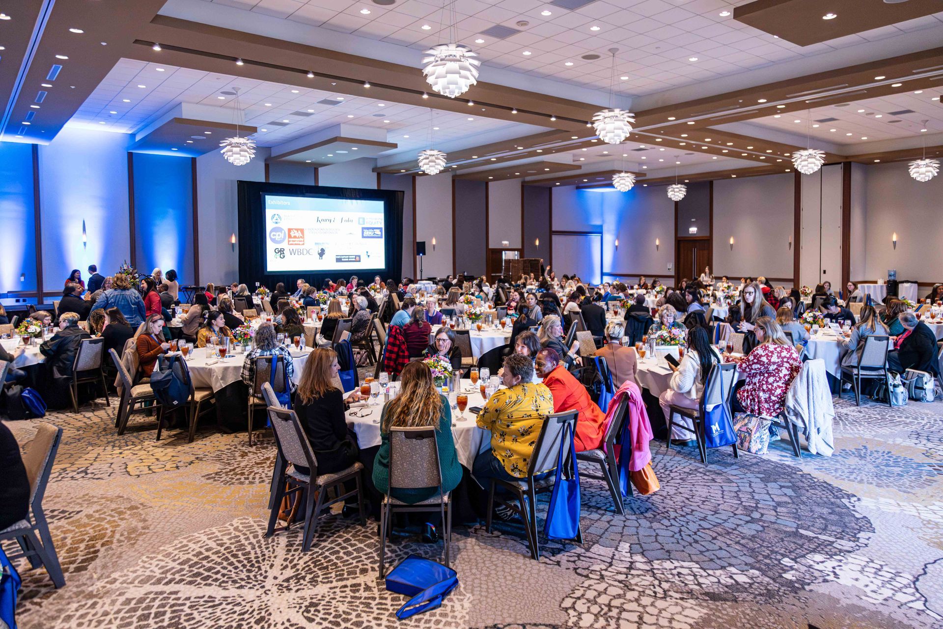 Large event in a ballroom; attendees seated at round tables. A screen displays presentation. Soft lighting, chandeliers.
