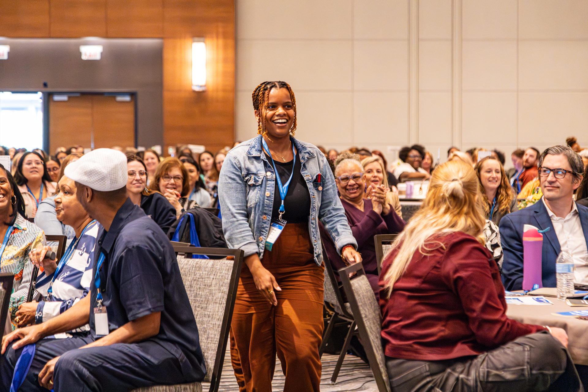 Woman in denim jacket smiles, walks between tables at a conference. Attendees applaud.