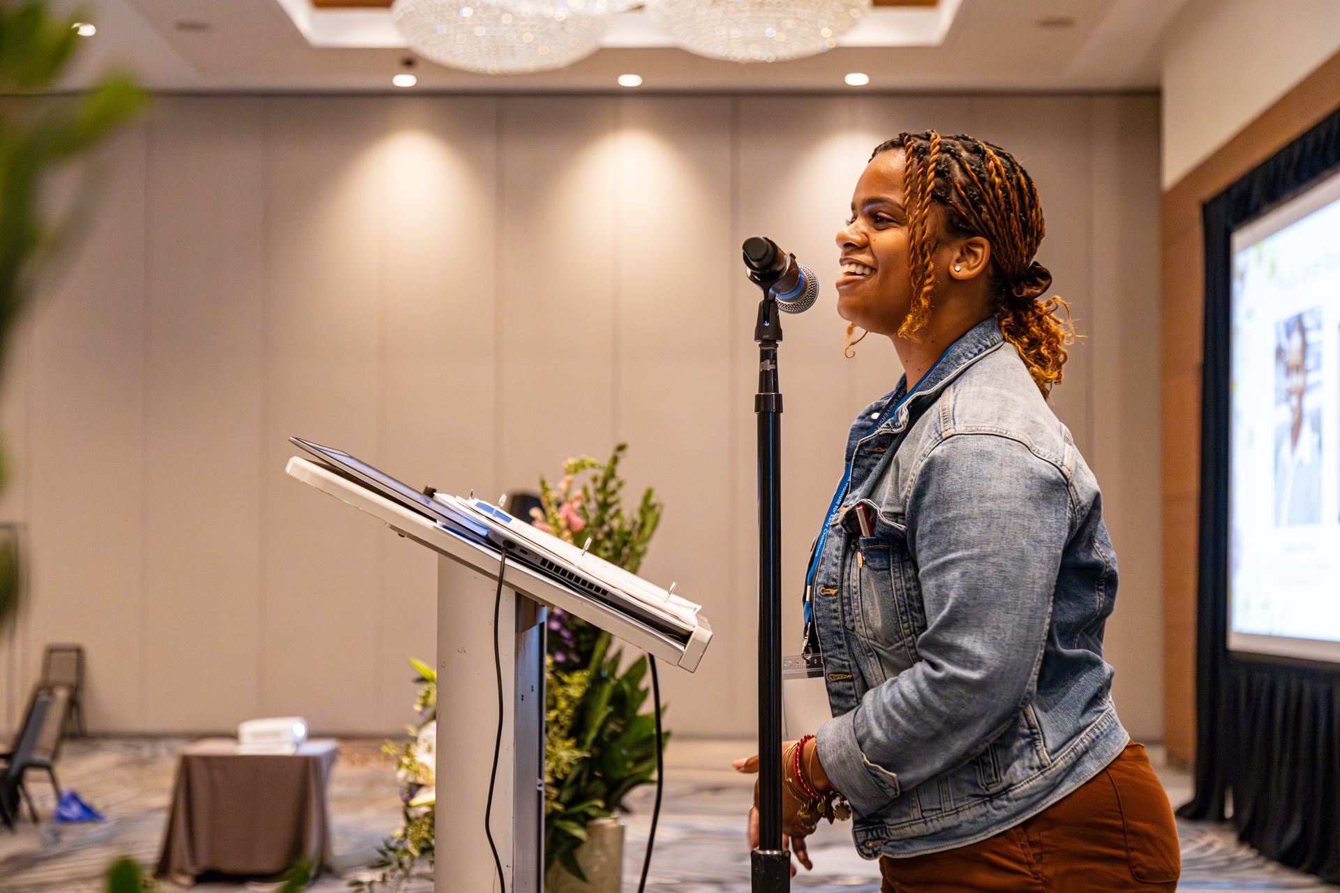 Woman speaking at a podium, microphone in front. She wears a denim jacket and smiles in a large room.