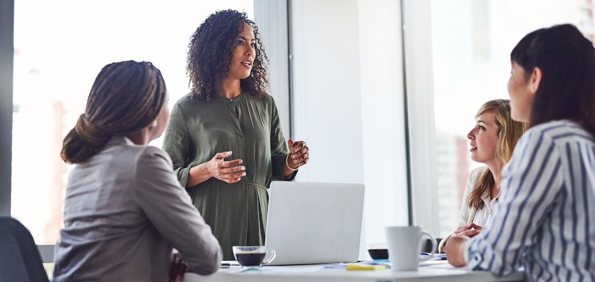 Four women in a meeting around a table, one woman is speaking, while others listen in an office.