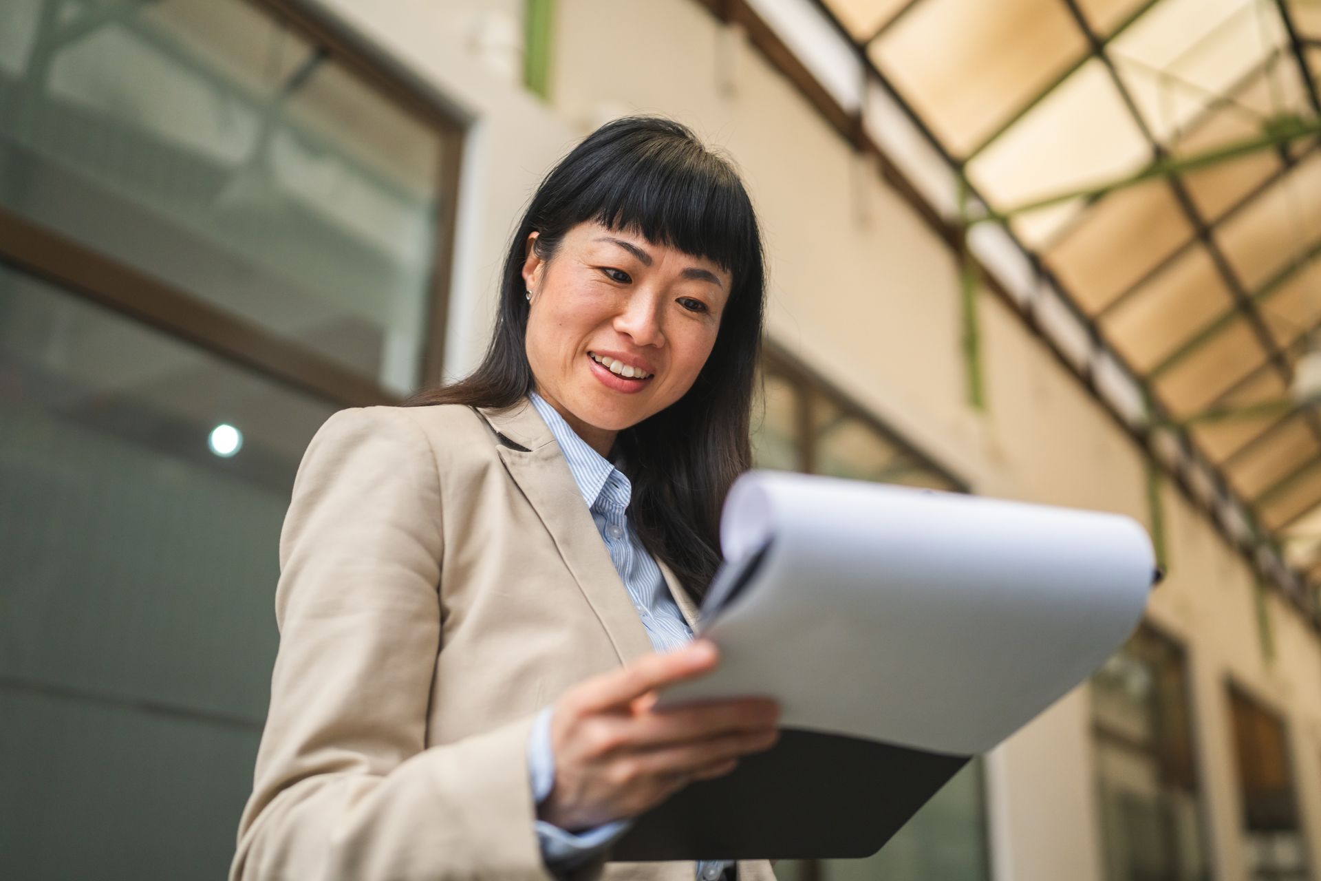 Smiling woman in beige blazer reviewing papers in a bright office hallway