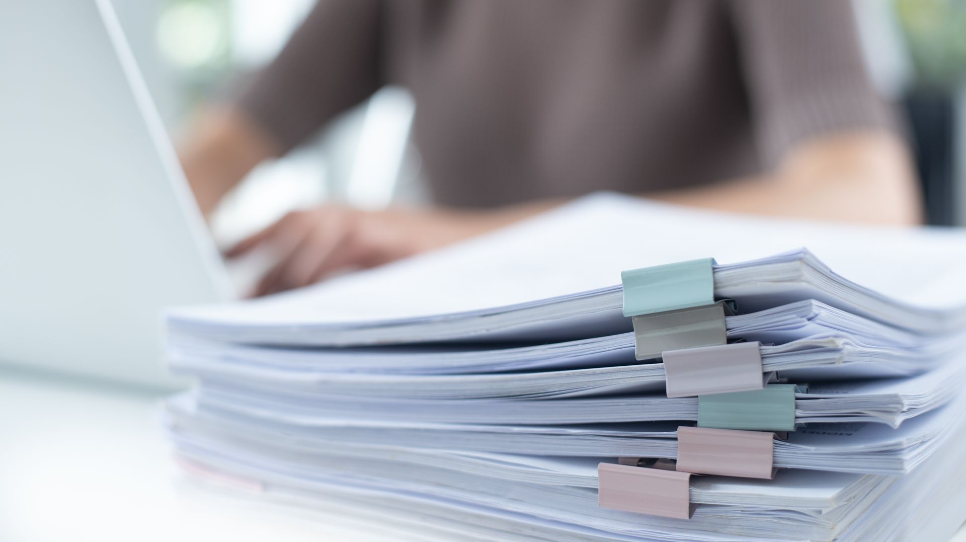 Pile of documents clipped together, with a person working on a laptop in the background.