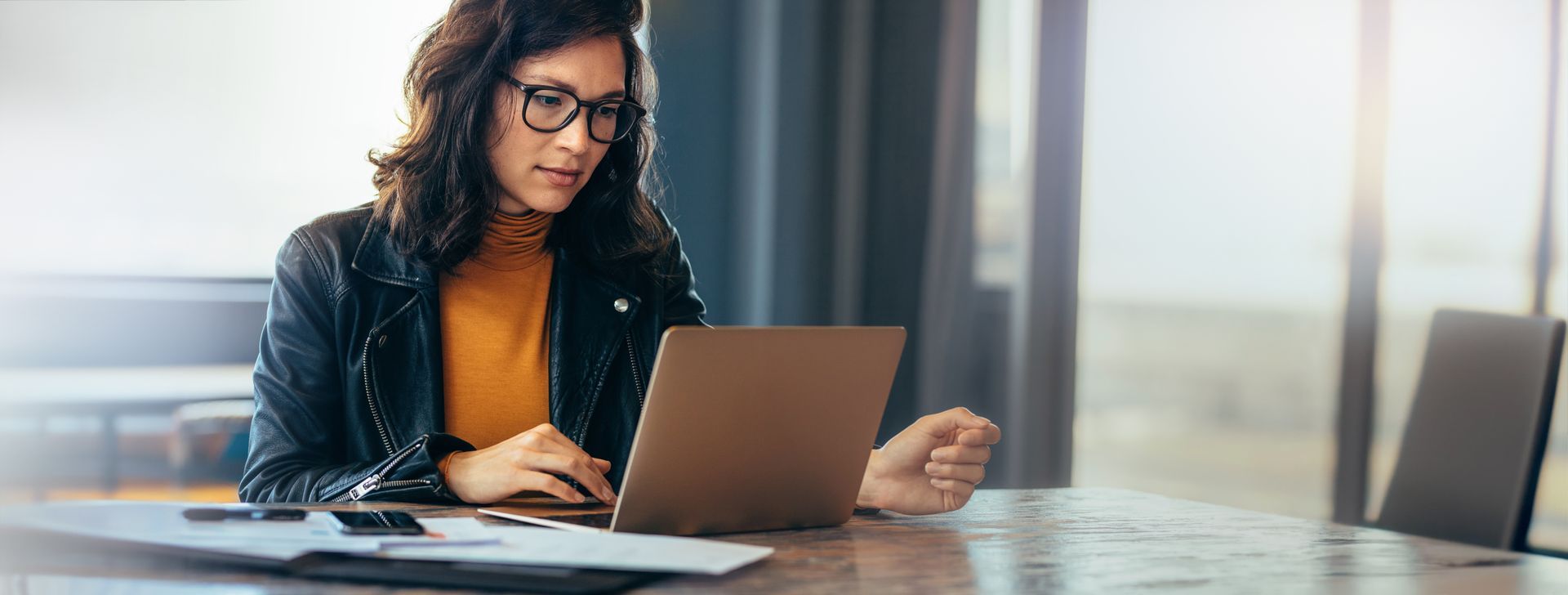 Person working on a laptop at a table in a bright office, with papers and a notebook nearby