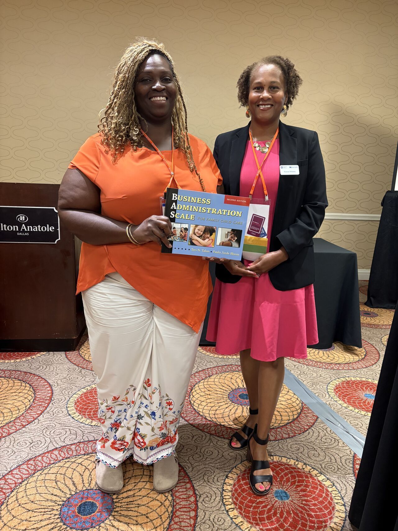 Two women smiling and holding a colorful booklet at an indoor event.