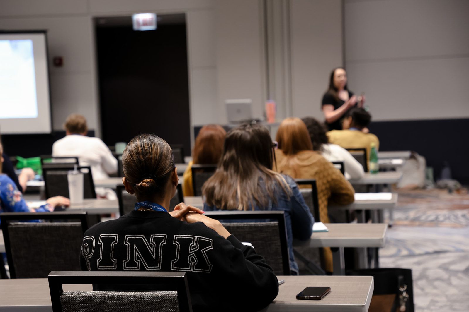 A woman presenting in a conference room with an audience seated at tables. Projector screen visible.
