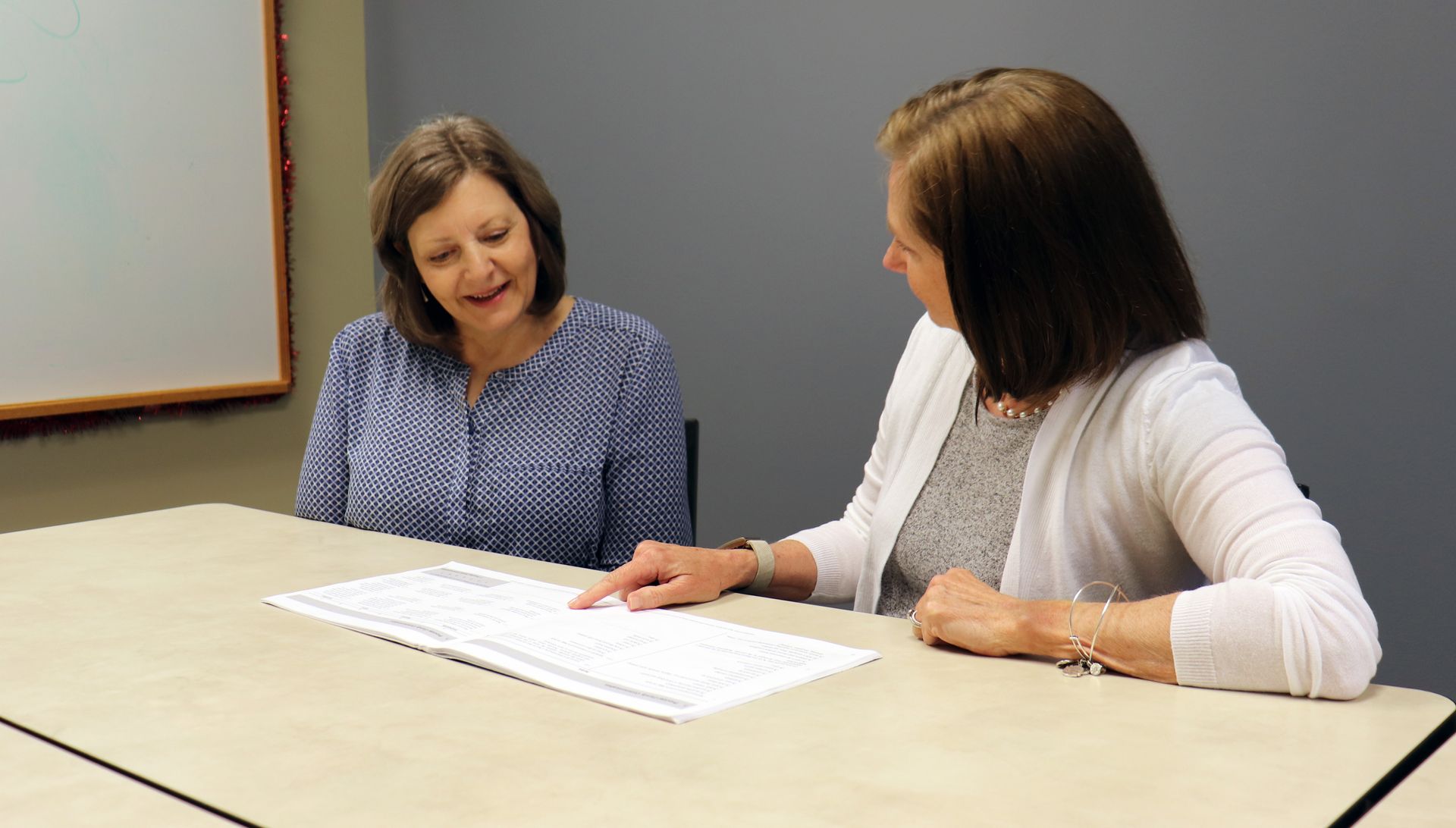 Two women seated at a table, looking at a document. One points to text; the other watches.