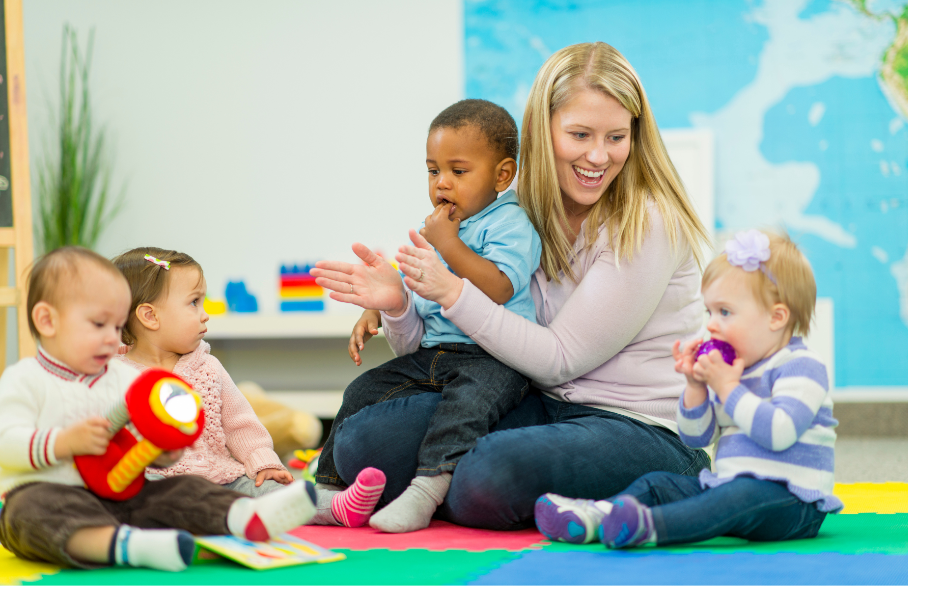 Teacher playing with children on a colorful classroom rug, holding a toddler and clapping with others nearby