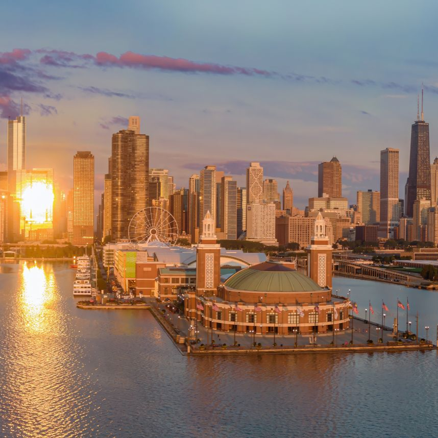 Chicago skyline at sunset; iconic Shedd Aquarium in foreground. Bright sun and buildings reflect on water.