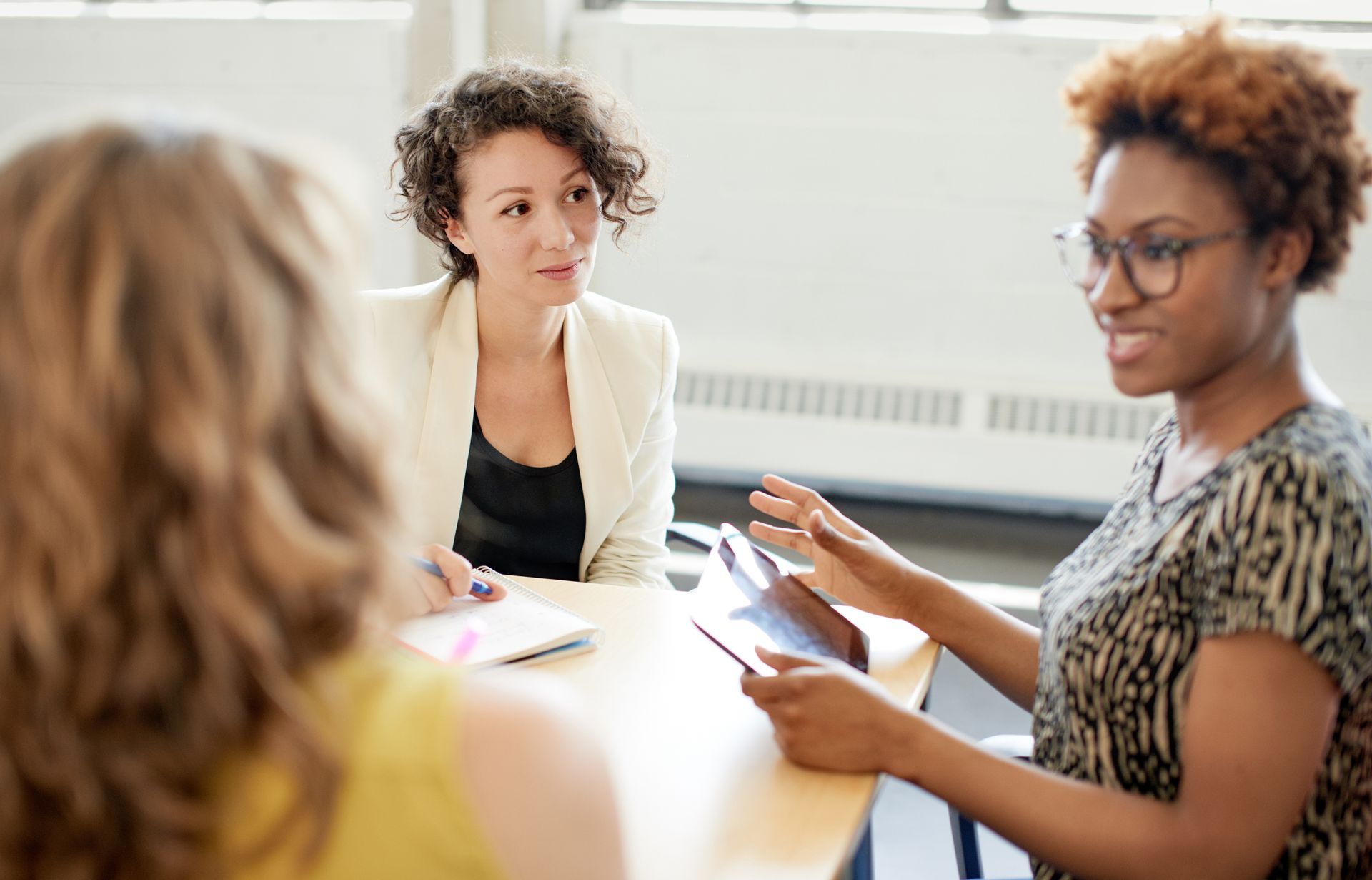 Three people in a meeting, one holding papers and speaking at a table in a bright office.