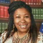 A woman with braids is smiling in front of a bookshelf.
