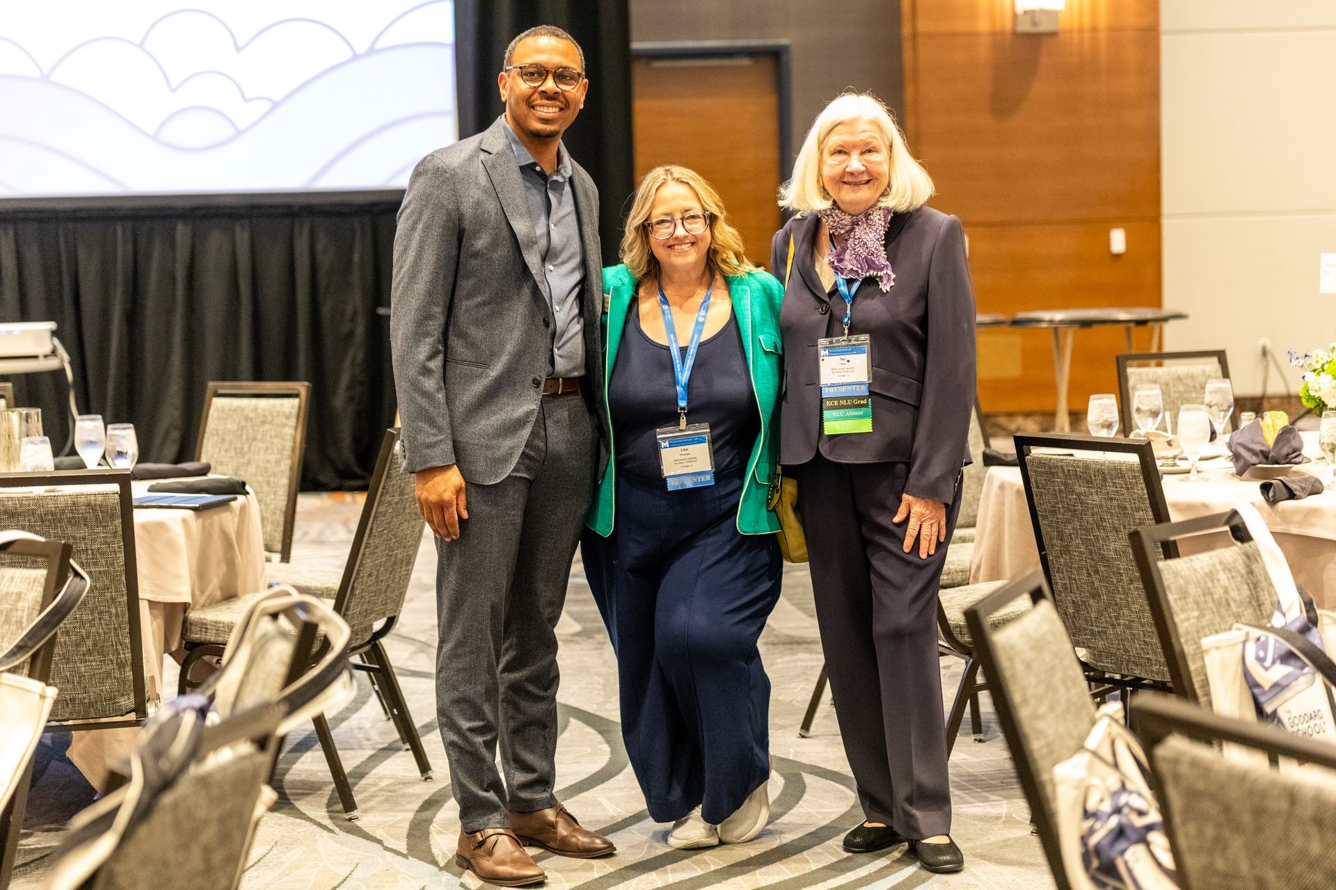 Three conference attendees posing and smiling in a meeting hall with banquet chairs and round tables.