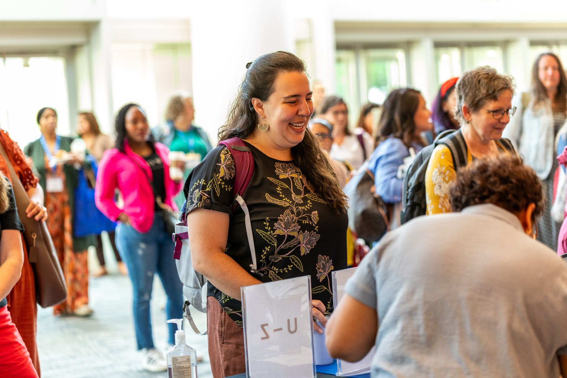 People mingling at an indoor event, with a smiling woman in black and gold in the foreground.